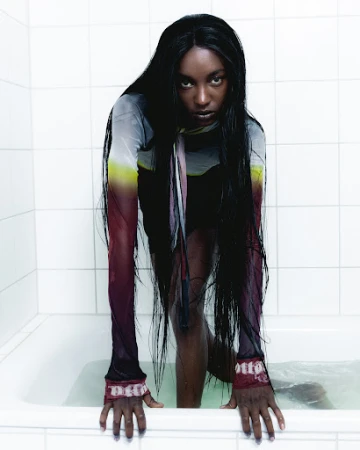 Dramatic portrait of a woman with long dark hair emerging from a bathtub filled with water, wearing a sheer printed top.