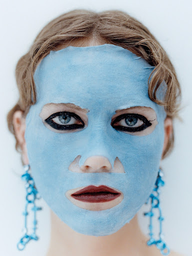 Close-up of a woman with a blue sheet face mask, dark eyeliner, and decorative blue earrings against a bright white background.