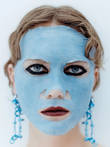 Close-up of a woman with a blue sheet face mask, dark eyeliner, and decorative blue earrings against a bright white background.