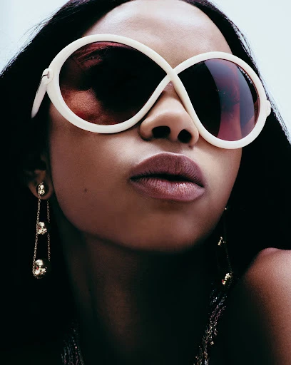 A close-up of a dark-skinned woman wearing oversized white-rimmed sunglasses and long gold earrings against a bright neutral background.