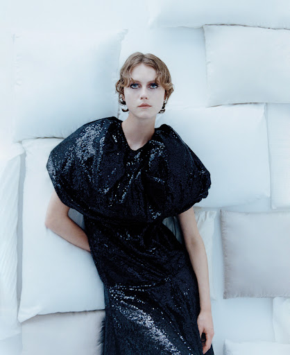 A fair-skinned young woman with short, wavy hair poses in a black sequin dress against a background of white pillows in a studio setting.