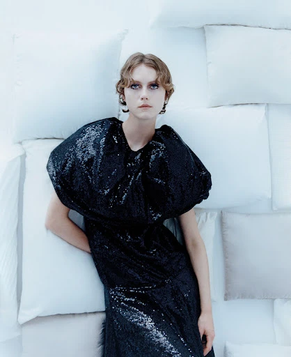 A fair-skinned young woman with short, wavy hair poses in a black sequin dress against a background of white pillows in a studio setting.