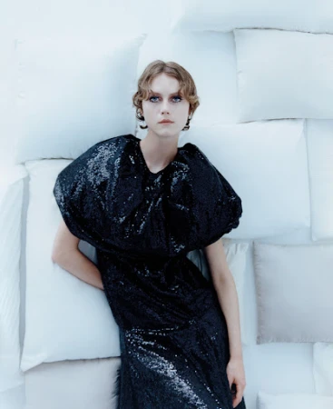 A fair-skinned young woman with short, wavy hair poses in a black sequin dress against a background of white pillows in a studio setting.
