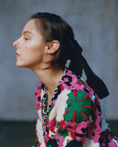 Profile view of a young woman with dark hair wearing a pink floral jacket with a black ribbon in her hair against a grey background.