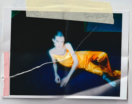 Fashion shot of a female model lying down in an orange outfit against a dark backdrop, with a torn photograph aesthetic and taped edges.