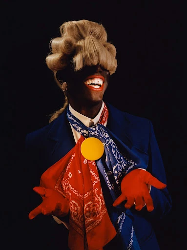 Person in a blue jacket and red gloves smiles with an elaborate blonde wig, gold pin, bandanas against a dark background in a portrait.