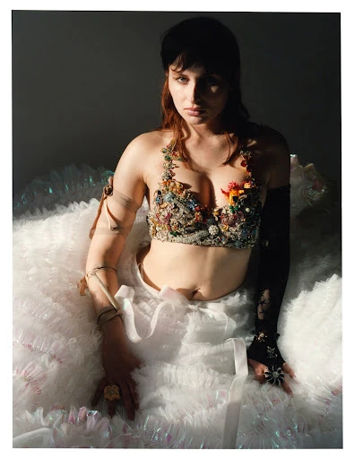 A woman poses in a studio wearing a jeweled bustier and a white ruffled skirt, with soft, diffused lighting and a dark backdrop.