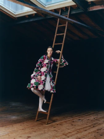 Model wearing a patterned floral coat and white stockings standing on a wooden ladder in a dark industrial loft with skylights