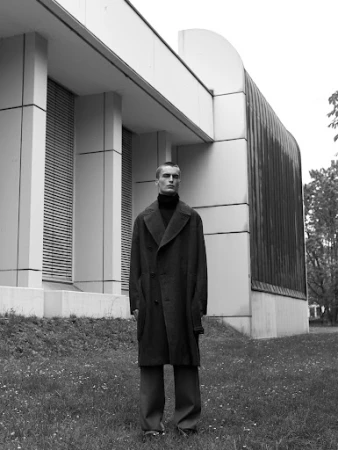 A young man wearing a dark overcoat and turtleneck standing on grass in front of a modern architectural building exterior.