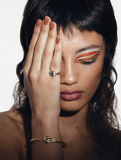 Close-up of a female model with creative makeup, showcasing a gold ring and bracelet against a bright white backdrop in a studio setting.