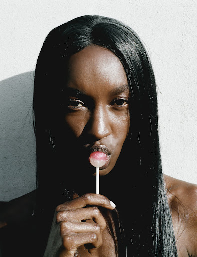 Close-up portrait of a Black woman with long, straight hair holding a lollipop to her lips against a bright white background.