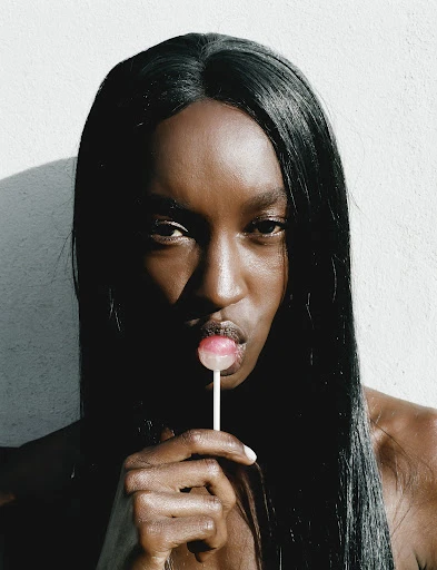 Close-up portrait of a Black woman with long, straight hair holding a lollipop to her lips against a bright white background.