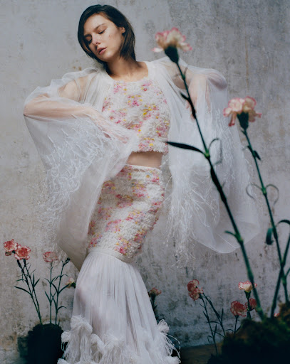 Young woman poses in a floral two-piece dress with flowing sleeves and a long skirt, surrounded by pink carnations against a textured wall.