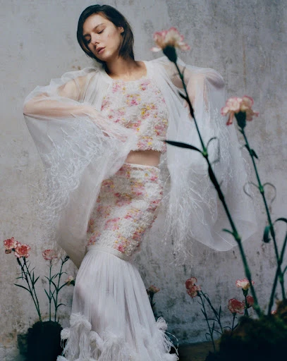 Young woman poses in a floral two-piece dress with flowing sleeves and a long skirt, surrounded by pink carnations against a textured wall.