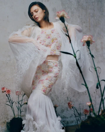 Young woman poses in a floral two-piece dress with flowing sleeves and a long skirt, surrounded by pink carnations against a textured wall.