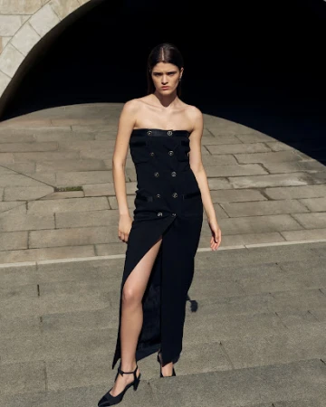 Model wearing a strapless black dress with front slit standing on stone stairs with arched architectural background.