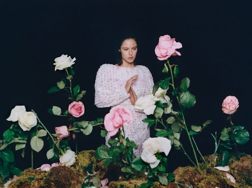 A woman poses in a dress surrounded by pink and white roses against a dark backdrop, creating a floral fashion portrait.