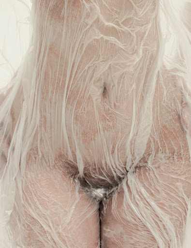 Close-up studio shot of a person draped in transparent plastic, showcasing the texture and form with neutral, soft lighting and skin tones.