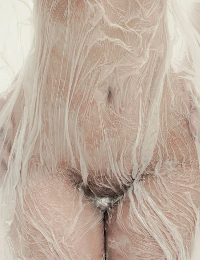 Close-up studio shot of a person draped in transparent plastic, showcasing the texture and form with neutral, soft lighting and skin tones.