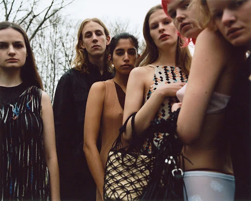 A group of six fashion models in an outdoor setting. Five adult females and one adult male pose for a fashion editorial shot.