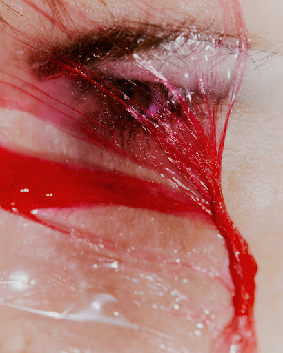 Close-up photograph of a person's face with red paint dripping around the eye and down the cheek, creating an abstract and dramatic effect.