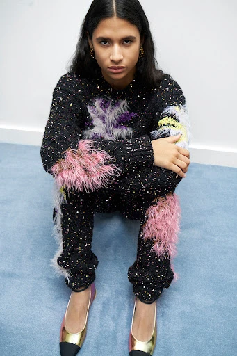 A woman in a black knitwear sweater and pants with pink feather accents poses indoors, seated on a blue surface with gold shoes.
