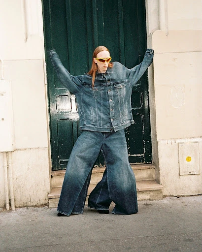 Person in oversized denim jacket and jeans strikes a pose on a city street with a green door in the background, wearing futuristic glasses.