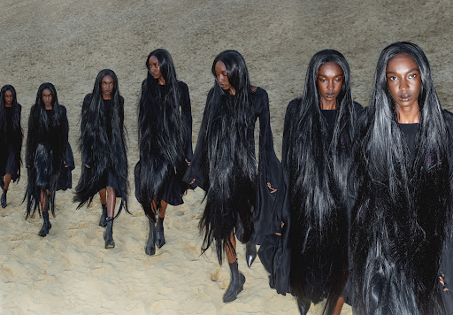 Runway models present avant-garde fashion during a fashion show, showcasing black dresses with long black hair on a sandy terrain.