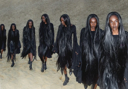 Runway models present avant-garde fashion during a fashion show, showcasing black dresses with long black hair on a sandy terrain.