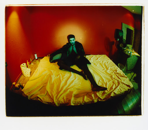 Person in dark clothing poses on a bright yellow bed in a red room; the person has makeup and dark hair and is staring forward.