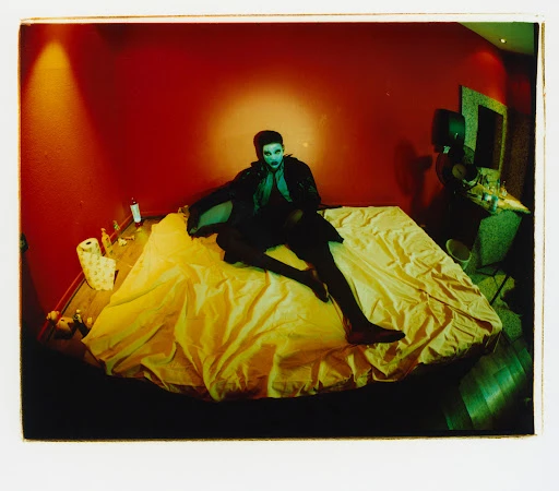 Person in dark clothing poses on a bright yellow bed in a red room; the person has makeup and dark hair and is staring forward.