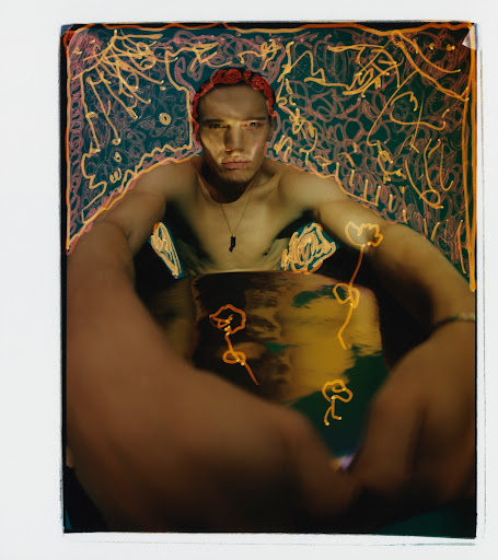 A shirtless man with a red headscarf sits in a container with water, framed by an illustrated floral and abstract background in warm light.