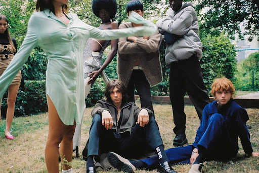 Group of young adults in stylish clothing gather on a grassy lawn, with trees and a building in the background, posing for a fashion editorial.
