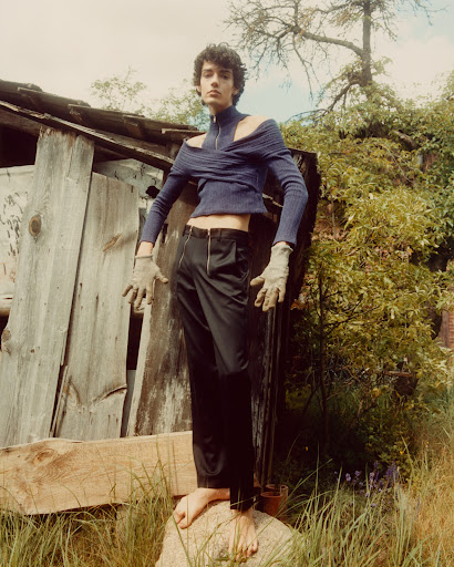Young adult stands on a rock in front of a shack, wearing a blue knit top and black pants, with natural outdoor light and muted tones.