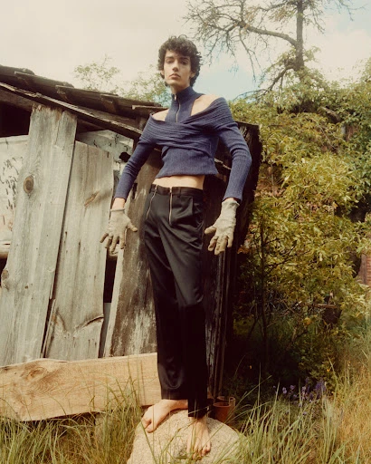 Young adult stands on a rock in front of a shack, wearing a blue knit top and black pants, with natural outdoor light and muted tones.