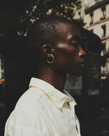 Profile portrait of a Black man wearing a light yellow collared shirt and silver hoop earring on a Paris street.