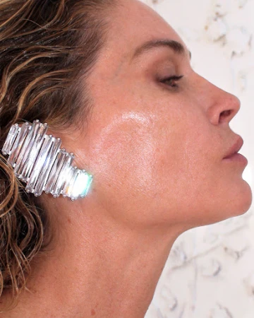 Close-up profile of a woman with wet hair wearing a crystal ear cuff and dewy makeup against a light background.