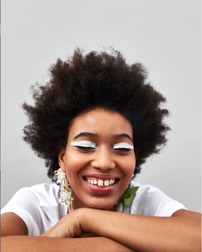 Close-up of smiling African-American woman with white eyeliner makeup, large earrings, wearing a white t-shirt, set against a gray background.