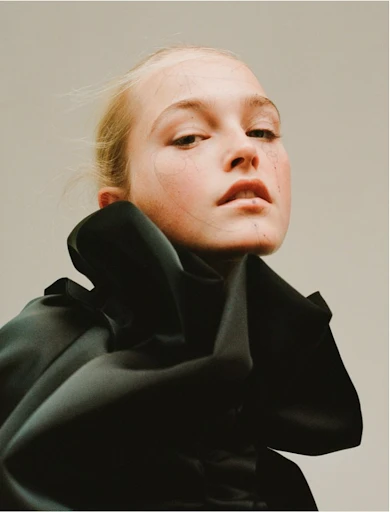 Close-up fashion portrait of a young adult female model with pale skin, wearing a dramatic black ruffled collar. Soft, diffused lighting.