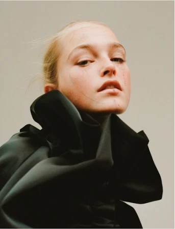 Close-up fashion portrait of a young adult female model with pale skin, wearing a dramatic black ruffled collar. Soft, diffused lighting.