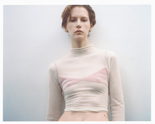 Woman in a sheer top stands against a white wall. Close-up shot in portrait orientation with natural light for a fashion editorial.