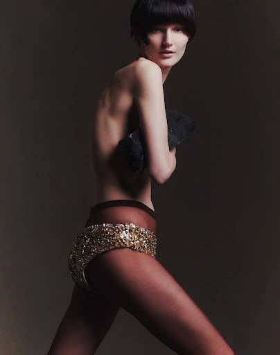 A slim woman with a dark bowl cut poses in bejeweled shorts with a dark top against a neutral backdrop in a fashion photograph.