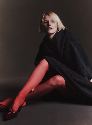 A woman poses in a black coat with red hosiery and high heels against a dark grey background in a fashion photography studio setting.