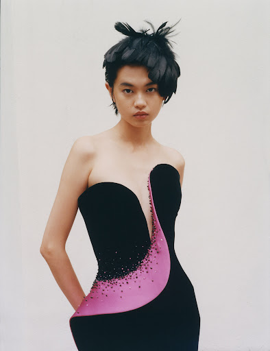 Woman in elegant black strapless dress with pink detail and feathers in hair poses against white wall in a medium shot.