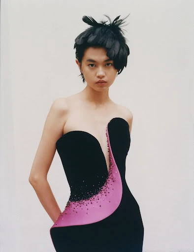 Woman in elegant black strapless dress with pink detail and feathers in hair poses against white wall in a medium shot.