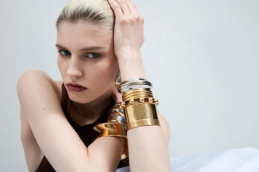 Close-up of a light-skinned woman posing with stacked gold bracelets, and a brown sleeveless top against a light background.