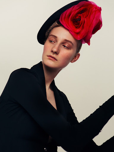 Woman in black jacket and hat with red flower detail gazes to the side in a fashion editorial portrait against an off-white backdrop.