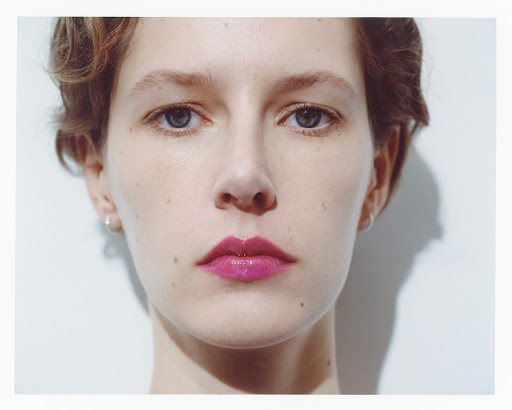 Close-up portrait of a woman with short hair and glossy magenta lipstick against a plain white wall