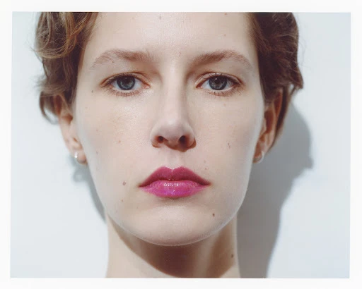 Close-up portrait of a woman with short hair and glossy magenta lipstick against a plain white wall