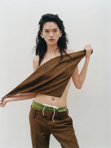 A young adult female poses in a studio, partially obscured by a brown cloth with logo, wearing matching trousers and statement earrings.